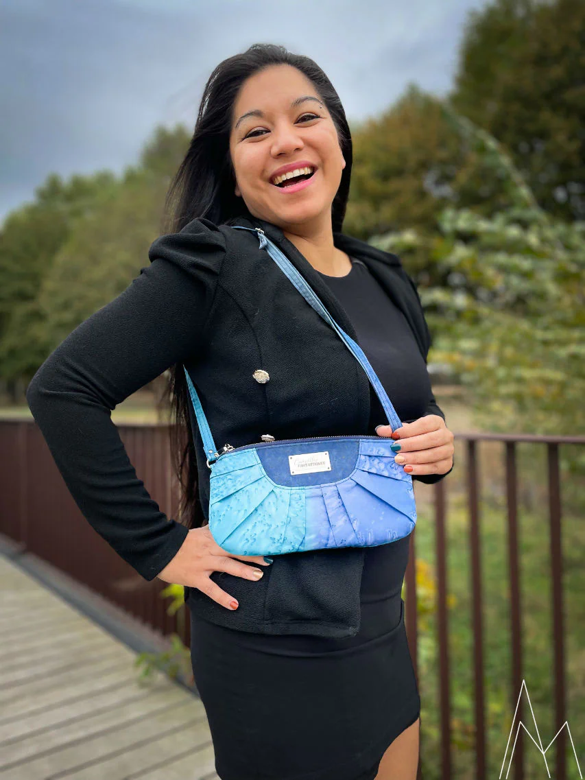 Photo of a young woman with tanned skin wearing a pleated shoulder bag in shades of blue, in an outdoor park during the day.