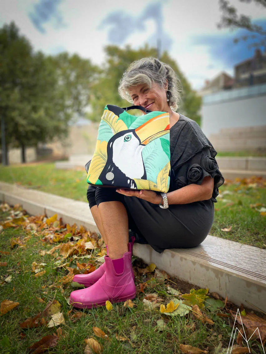 Photo of a young white woman with blonde hair wearing a white backpack with a rainbow pattern resembling a unicorn, in an outdoor park during the day.