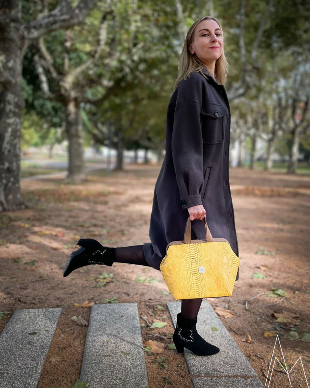 Photo of a young white blonde woman carrying a yellow and brown handbag, in an outdoor park during the day.