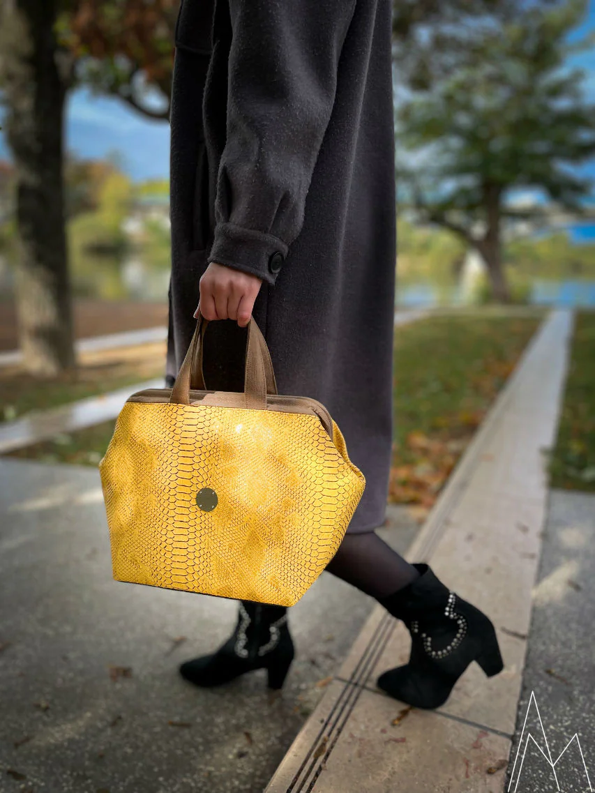 Photo of a young white blonde woman carrying a yellow and brown handbag, in an outdoor park during the day.