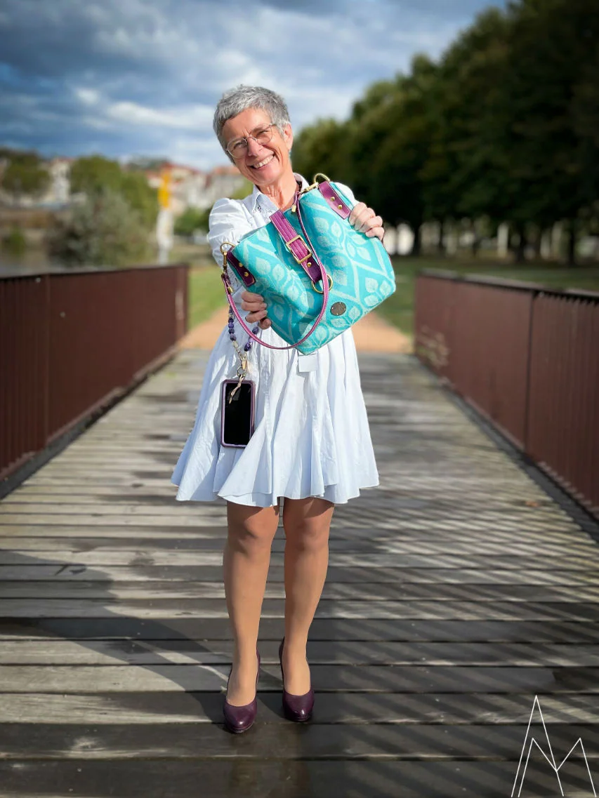 Photo of a mature white woman carrying a turquoise and plum shoulder bag, in an outdoor park, during the day.