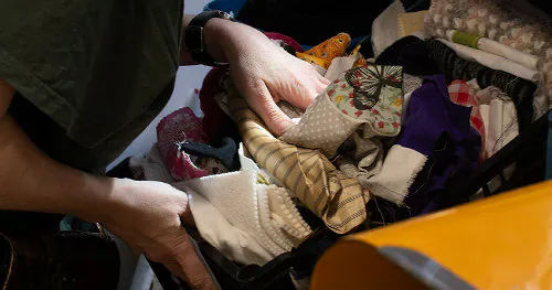 Hands sorting through a crate full of mixed fabric offcuts — wool, velvet, cotton, silk — ready to be repurposed.