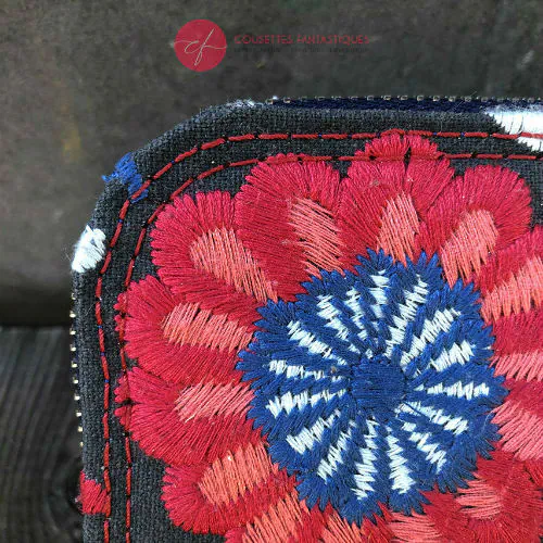 Un petit porte-cartes et porte-monnaie zippé en tissu gris brodé de motifs floraux scandinaves bleus, rouges et blancs.