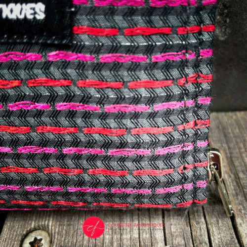 Un petit porte-cartes et porte-monnaie zippé en tissu gris brodé de lignes roses et rouges brillantes, avec un intérieur en toile gris mat.