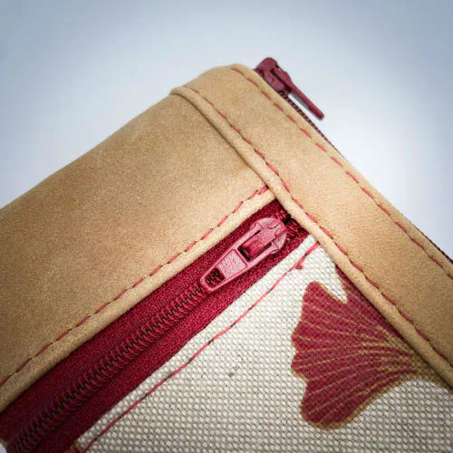 Un mini portemonnaie zippé fait de cuir de couleur camel et d'une toile en polycoton avec un motif de feuilles de Ginkgo Biloba rouges.