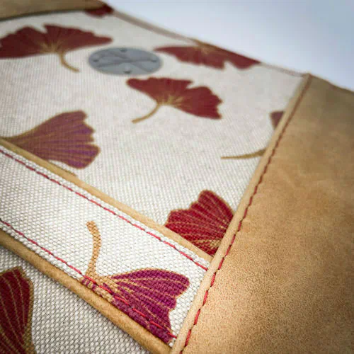Une pochette zippée cousue dans un cuir de couleur camel et une toile en polycoton avec un motif de feuilles de Ginkgo Biloba rouges.