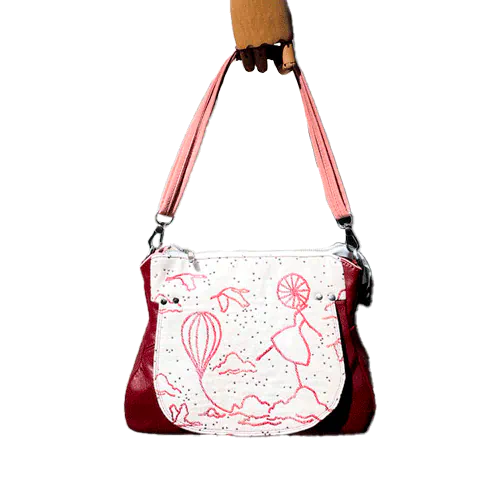 Un sac d'épaule fait avec du tissu d'écharpe écru, framboise, orange et corail, au motif poétique de montgolfières et de valseuses, et du cuir prune.