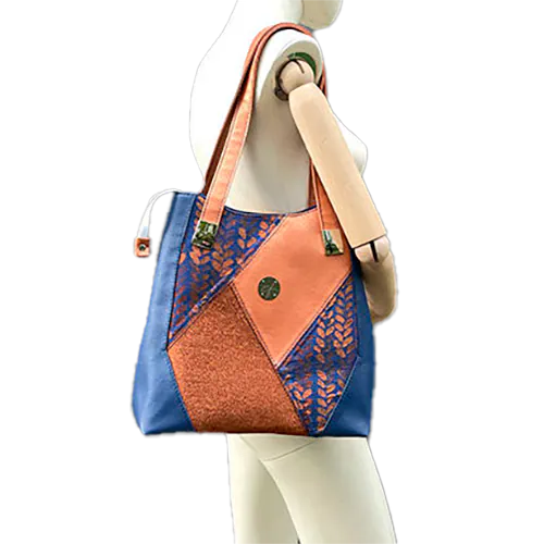 Un sac d'épaule composé d'un patchwork de différents matériaux : similicuir bleu royal, similicuir doré, tissu brillant bronze, et tissu wrap bleu et bronze avec un motif de petites gouttes.